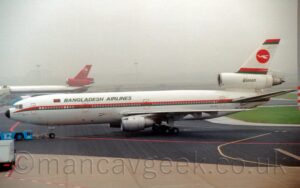 Side view of a 3 engined jet airliner, facing to the left but being pushed back to the right by a low-slung blue tug attached to the nose-wheel by a long grey pole. The plane is mostly white, with a red and green stripe running along the body, covering the passenger cabin windows, with green "Bangladesh Airlines" titles on the upper forward fuselage, next to a green and red Bangladeshi flag. The black registration "S2-ACO" on the lower rear fuselage, with the text "DC-10-30" under the horizontal stabiliser. There are green "Biman" titles on the side of the white centre engine pod, running through the white tail, with red and green stripes at the top and the bottom of the tail, with a red circle with a stylised white bird in the middle. Black and grey apron, marked with yellow and red stripes, fill the foreground, with patches of green grass on the right of the frame. Another 3 engined jet airliner is parked facing to the left in the background, this one silver and black with a red tail, parked nose0in to a terminal building, the rest of the background swallowed up by hazy grey sky.
