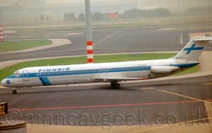 Side view of a twin engined jet airliner taxing from right to left, following a yellow line along a black taxiway. The plane is mostly white, with a blue stripe running along the body, covering the passenger cabin windows. There are blue "FinnAir" titles on the upper forward fuselage, with a diagonal blue band under the lower forward fuselage, and the blue registration "OH-LYU" on the upper rear fuselage, just in front of the white engine pods mounted on the sides of the rear fuselage. Black taxiway and grey apron fills the foreground, with islands of green grass in the background surrounded by grey concrete borders in a sea of black taxiway, with a tall, grey lighting pole with a red and white banded base immediately behind this plane, rising up out of the top of the frame. Trees and buildings in the distance are swallowed up in gey mist.