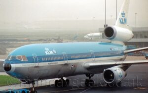 Side view of a 3-engined jet airliner being towed from right to left along a black taxiway by an almost-out-of-frame blue tug. The plane is light blue on top, with a darker blue stripe running along the body, covering the passenger cabin windows, above a thinner white stripe, and a grey lower half. There are white "KLM" titles under a stylised white crown on the upper forward fuselage, repeated in a larger form in light blue on the white tail, with a smaller version with a light blue crown and dark blue text on the sides of the white and grey engine pods under the wings. The white registration "PH-DTC" is on the upper rear fuselage, just aft of small EU and Dutch flags There is light blue text "Frederic Francois Chopin" on the white stripe, just below the cockpit windows, with dark blue text "Royal Dutch Airlines on the same stripe below the large "KLM" logo. Black taxiway fills the foreground, with a grey terminal building stretching half-way into frame from the right, and a building site on the taxiway on the left of the background, with a white jet airliner moving out from behind the terminal building, heading past the building site. Another 3-engined jet airliner is barely visible on a runway in the distance, almost swallowed up by mist, with a white building behind it, with hazy grey clouds filling the rest of the frame.