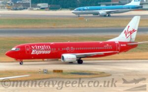 Side view of a twin engined jet airliner taxiing from right to left along a grey taxiway. The plane is mostly red, with large, white "Virgin Express" on the forward fuselage, above and below the forward passenger cabin windows. The white registration "OO-VED" is on the side of the rear fuselage, just aft of the rear door, with a small, black, yellow, and red Belgian flag and the white text "Boeing 737-400" just in front of the door. The tail is white, with red, seemingly hand-written "Virgin" titles in the middle. A couple of grey taxiways meet in the bottom right corner, separated on the left by a strip of grass. More grass in the background separates this taxiway from a black runway, with more grass beyond that separating another grey taxiway. A similar type of plane with a blue, dark blue, and white livery is taxiing from right to left in the top right corner.
