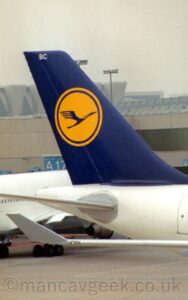 Vertically-oriented close-up of the white tail-cone and blue tail of a jet airliner taxiing from left to right. There is a yellow circle, outlined in yellow, in the middle of the tail, itself containing a highly stylised image of a blue flying bird, with the white letters "BC" right at the top. The end of the wings are visible in the foreground, the white, up-turned wing-tip tilted slightly outwards. Another white jet airliner is parked in the background on the left, with a grey terminal building stretching across the frame beyond that, under flat grey sky.