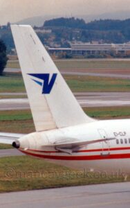 Vertically-oriented close-up of the rear fuselage and tail of a jet airliner taxiing from left to right along a grey taxiway. The plane is mostly white, with a pair of red stripes running along the body, and the black registration "EI-CLP" on the upper rear fuselage, above the rear-most passenger cabin windows. There is a stylised blue letter "V" on the tail. Grey taxiways and a runway run from side to side in the background, lined with wide strips of green grass, with grey buildings in the background, in front of low hills covered in trees, under hazy grey sky.
