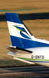 Vertically-oriented close-up of the tail-cone and tail of a propellor-engined airliner taxiing from left to right. The plane is white, with a thin green and blue pinstripe running along the body, under the black registration "G-GNTD". The tail is blue, with a diagonal white band wrapped around the middle, with a stylised, reversed, lower-case black letter "b" with green, white, and blue high-lights in the middle. A black taxiway runs across the bottom of the frame in the background, with yellowing grass filling the rest.
