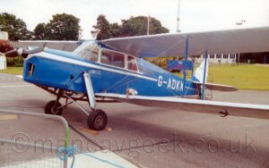 Side view of a single-engined bi-planes parked facing to the left on black asphalt, with wooden block in front of and behind the left main wheel, preventing it from moving. The plane is largely blue, with a white stripe running along the body from the propellor, overlaid with the white registration "G-ADKK" on the side of the rear fuselage. The windows of the enclosed, 4-seat cockpit are outlined in white, and the wings are also white. Black asphalt fills the foreground, with green grass running across the background, with trees on the left and a white building on the right, under bright but hazy sky.
