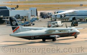 Side view of a high-winged, twin propellor-engined airliner taxiing from left to right and slightly away from the camera. The plane has a grey top half with a dark blue, almost black bottom half, with a thin red stripe running along the body, ending in a half arrowhead under the cockpit, behind grey "Operated by CityFlyer" text. The grey registration "G-BVTJ" is on the lower rear fuselage. The top half of the tail is dark blue, and carries a grey Royal Crest, while the bottom half is grey, with a red and dark blue triangle at the rear. Grey concrete apron fills the foreground and a large chunk of the background, with airport vehicles scattered around, and a white, high-winged, 4 engined jet airliner parked in the upper right corner, and the end of a black terminal building with a couple of empty, white air bridges on the upper left.