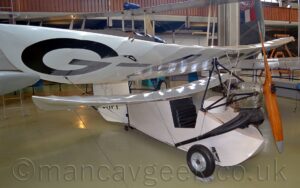 Front-side view of a single-engined, single-seat light aircraft, with a pair of staggered wings, on display facing to the right in a museum. The plane is white, with a an open cockpit in the nose behind the exposed engine. A large, white, ribbed wing is suspended over the cockpit, with part of the registration "G-A" visible under the right side. A smaller, white wing is mounted on the top of the fuselage immediately behind the cockpit. The white tail has the text "BHP1 at the bottom, and a line drawing of a dog, both partially obscured by the wings. There is no horizontal stabiliser. A creamy-grey floor fills the foreground and a large chunk of the background. A grey bi-plane fills most of the rest of the background on the left, with the grey tail of a larger aircraft, with the red number "80", outlined in white, and a red, white, and blue finflash on the side, on the right. A walkway with a brown wooden balustrade stretches across the top of the frame.