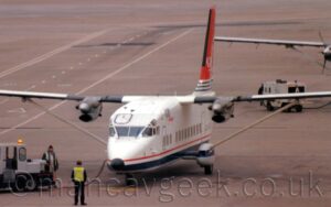 Front of a box-like, high-winged, twin propellor-engined airliner parked on a grey concrete apron area, facing slightly off to the left of the camera. The plane is largely white, with a thin red pin-stripe and a thicker blue stripe running along the bottom of the body with a grey belly. There are blue "Gill" and red "Airways" titles on the upper forward fuselage, and the black registration "G-DASI" just visible on the lower rear fuselage. The tail is largely red, with white "Gill Airways" titles across the middle, and a series of thin white and blue stripes at the bottom. White, blue, and grey sponsons housing the main landing gear stick out from the sides of the bottom of the fuselage, with long support struts coming from the tops, stretching out to support the outer panels of the long, thin wings. Two men wearing hi-viz vests are standing in the bottom left corner of the frame, next to a small grey and dark blue truck, connected to the planes nose by red and black electrical cables. Grey concrete apron fills the rest of the frame, with a roadway marked out with white lines on the left, and the wing of a similar plane sticking in to frame on the right.