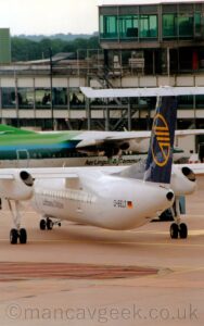 Vertically-oriented rear view of a high-winged, twin propellor-engined airliner, facing to the left but being pushed back to the right by a low-slung blue tug attached to the nose-wheel by a long white pole.The plane is largely white, with dark blue "Lufthansa CityLine" titles on the lower forward fuselage, and the black registration "D-BELT" next to a small German flag on the side of the rear fuselage. The tail is dark blue, with the yellow outline of a circle, containing a highly-stylised, angular image of a yellow flying bird, in the middle of the tail. Grey concrete apron fills the foreground, with a white and grey terminal building stretching across the background, with people inside visible through the large widows, and a green and white airliner parked nose-in on the left. Green trees under bright but hazy sky fills the top left corner of the frame.