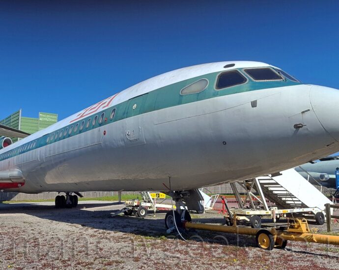 Low-down close-up of the nose and cockpit of a three-engined jet airliner, parked facing to the right, with the rest of the fuselage and tail visible on the left of the frame. The plane is white on the top, with a grey lower half, and a dark blue stripe, faded to green, running along the body, covering the passenger cabin windows. There are white "BEA" titles with large red drop-shadows on the upper forward fuselage. The black registration "G-AWZK" is on the lower rear fuselage, just forward of the side-mounted engines. The centre-engine intake at the bottom of the front of the tail has a red baffle inside, with white text "Trident Three" on the side of the faded green intake, and a quarter of a British flag on the side of the tail. A long yellow pole is attached to the plane's nose-wheel, running out of the lower right of the frame. A set of white air-stairs is parked against the forward door on the other side of the plane. Grey gravel fills the bottom of the frame, with another, military grey aeroplane visible under this plane's nose. Trees and a large, green metal box tower over the plane on the left of the frame. Vivid blue sky fills the rest of the frame.
