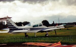 Rear side view of a twin propellor-engined light aircraft parked facing to the right and away from the camera. The plane is mostly white, with two black stripes either side of a blue stripe running along the body, getting wider as it moves rearwards, with additional, short, black stripes on the rear of the tail, with broadly similar stripes on the engine pods on the wings. The white registration "N453C" is overlaid on the stripes on the sides of the rear fuselage. Green grass fills the foreground, with red and yellow stripes sheets of metal in the bottom right corner. Another light aircraft is in the background on the right, with trees running along the skyline in the distance, under grey and white cloud.