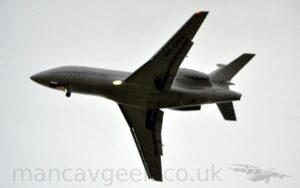 View from below of a 3 engined BizJet flying from right to left at a low altitude, with flaps extended and the undercarriage lowered, suggesting it is about to land. The plane is mostly white, with a black stripe swooping it's way along the lower fuselage, as well as along the tail. The black registration "9H-GRS" is on the sides of the engine pods mounted on the sides of the rear fuselage, as well as under the left wing. A bright light is shining in the wing root, slightly blowing out that part of the frame, as well as another light on the nose-wheel. Hazy grey sky fills the rest of the frame.