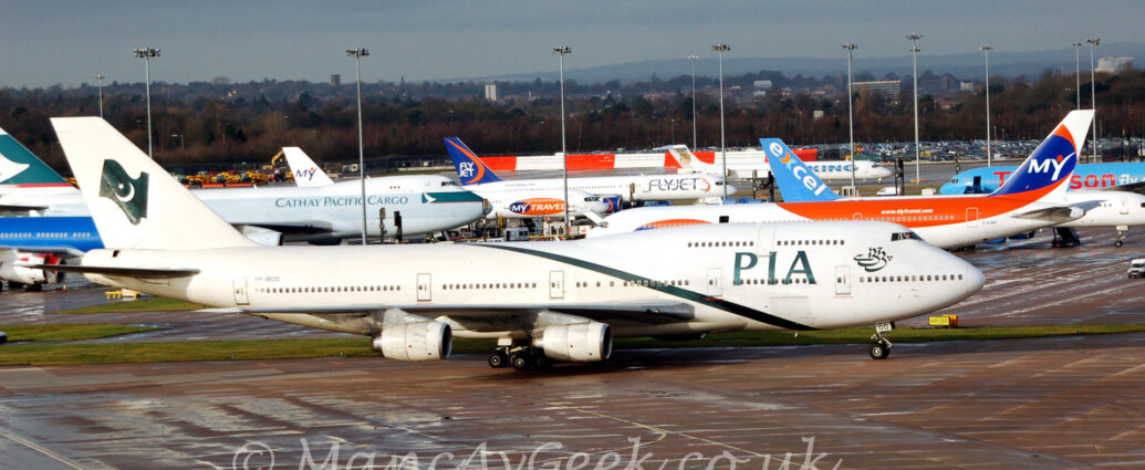 Side view of a very large, 4 engined jet airliner taxiing from left to right along a black taxiway. The plane is mostly white, with a thin, diagonal band running from under the forward fuselage, just aft of the nose-wheel, to over the top of the centre-fuselage, just aft of the wings, and large, green "PIA" titles on the side of the forward fuselage, between the upper and lower passenger decks, partially covering some of the lower deck windows. There is some ornate Arabic calligraphy on the upper forward fuselage, just below and aft of the cockpit, which takes the name of the airline in Arabic and turns it into a piece of art. There is a waving green flag on the white tail, with a white star and crescent moon in the centre. Damp, black apron fills the foreground, with a multitude of airliners, mostly white with orange, blue, green, and red highlights, parked in the background, mostly facing to the right, but a couple on the right of the frame are facing left. Tall lighting poles are dotted around the planes in the background, reaching up to the sky. Trees in the distance give way to a rolling landscape, with buildings and more trees leading off to the horizon, meeting cloudy grey skies.