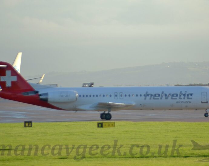 Side view of a white, twin engined jet airliner with the engines mounted on the sides of the rear fuselage, taxiing from left to right along a grey taxiway. There are large, grey "Helvetic" titles on the side of the forward fuselage, covering the passenger cabin windows, above smaller grey "Airways" titles. The grey registration "HB-JVF" is on the sides of the white engine pods mounted on the sides of the rear fuselage. A diagonal red band wraps around the tail and rear fuselage, with a large white cross in the middle of the tail. Lush green grass fills the foreground, while there is a blue and white airliner trying to sneak out of frame on the left of the background, and rolling hills in the distance, under bright but hazy sky.