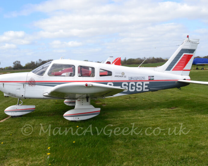 Side view of a single-engined light aircraft parked facing to the left on grass. The plane is mostly white, with a grey band wrapped around the lower rear fuselage, and a thin red pinstripe running along the middle of the body. The white registration "G-SGSE" is on the side of the lower rear fuselage, under the small black text "Archer II" and a block of text in a flowing Arabic script, which has a small Palestinian flag overlaid with the words "Free Palestine" through the middle next to it. A red stripe and a pair of grey stripes run through the middle of the spats on the tri-cycle under-carriage. Green grass fills the foreground, with several more light aircraft parked on more grass in the background. Trees line the airfield perimeter, meeting blue sky with patches of fluffy cloud.