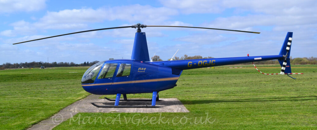 Side view of a blue helicopter parked on a concrete landing pad, surrounded by green grass. There is a gold stripe running along the body, with large gold text "R44" above smaller gold text "Raven II" just aft of the cockpit doors. The gold Registration "G-OGJC" sis on the long, blue tail-boom, which has a 2-bladed, black and white stripes rotor at the end, above a red and white striped pole attached to the underside of the boom. A tall blue fairing is wrapped around the main rotor mast. Green grass in the background stretches off into the distance, with trees on the horizon meeting blue sky with fluffs of white cloud.