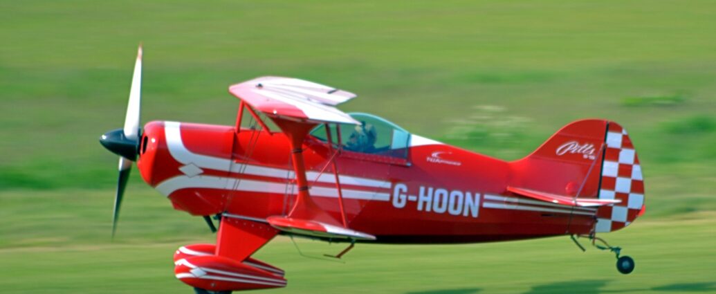 Side view of a small, red, single-seat bi-plane flying from right to left at a very low altitude, just a couple of metres above the ground, quite possibly in the middle of taking off. The plane is almost entirely red, with a pair of white stripes running along the body, looping in from above and under the engine cowling in the nose, overlaid with the white registration "G-HOON" on the side of the rear fuselage. A similar design, minus the registration, is on the spats covering the main landing gear at the front of the plane. The front half of the tail is the same bright red, with white text "Pitts S-1S" at the top, while the rear is covered in a red and white chequerboard pattern. The wings are red, with similar but thicker white lines running from front to back, and along the width. Green grass fill the rest of the frame, blurred into a smear, with the plane casting a strong shadow on the airfield below it, suggesting the sun is fairly low down on the left of the frame.