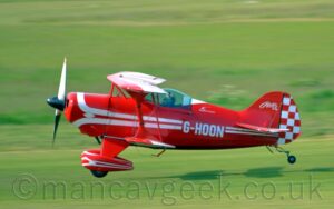 Side view of a small, red, single-seat bi-plane flying from right to left at a very low altitude, just a couple of metres above the ground, quite possibly in the middle of taking off. The plane is almost entirely red, with a pair of white stripes running along the body, looping in from above and under the engine cowling in the nose, overlaid with the white registration "G-HOON" on the side of the rear fuselage. A similar design, minus the registration, is on the spats covering the main landing gear at the front of the plane. The front half of the tail is the same bright red, with white text "Pitts S-1S" at the top, while the rear is covered in a red and white chequerboard pattern. The wings are red, with similar but thicker white lines running from front to back, and along the width. Green grass fill the rest of the frame, blurred into a smear, with the plane casting a strong shadow on the airfield below it, suggesting the sun is fairly low down on the left of the frame.