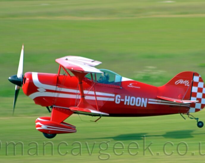 Side view of a small, red, single-seat bi-plane flying from right to left at a very low altitude, just a couple of metres above the ground, quite possibly in the middle of taking off. The plane is almost entirely red, with a pair of white stripes running along the body, looping in from above and under the engine cowling in the nose, overlaid with the white registration "G-HOON" on the side of the rear fuselage. A similar design, minus the registration, is on the spats covering the main landing gear at the front of the plane. The front half of the tail is the same bright red, with white text "Pitts S-1S" at the top, while the rear is covered in a red and white chequerboard pattern. The wings are red, with similar but thicker white lines running from front to back, and along the width. Green grass fill the rest of the frame, blurred into a smear, with the plane casting a strong shadow on the airfield below it, suggesting the sun is fairly low down on the left of the frame.