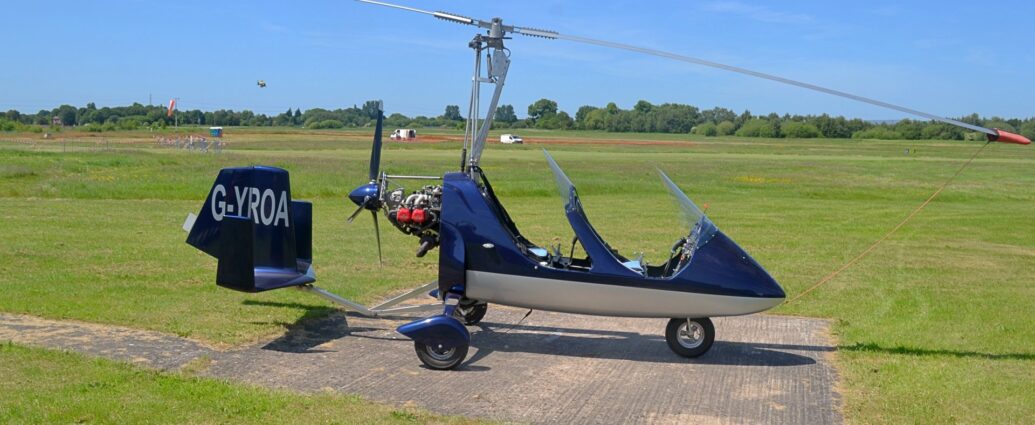 Side view of a two-seat, open-cockpit auto-gyro parked facing to the right on a concrete pad surrounded by grass. The plane's body is mostly blue, with a white belly, with blue covers over the rear tricycle wheels, and a metal frame extending backwards from under the rear-mounted engine, holding the blue tai, as well as the blue horizontal stabiliser, with blue, vertical end plates on the ends of the stabiliser. The white registration "G-YROA" is on the tail. Another metal frame rises up from behind the rear cockpit, holding the main rotor. A red sleeve covers the end of the main rotor above the front of the plane, attached to the nose by a long red cord. Green grass fills the foreground and most of the background, with a couple of white vans on the grass in the distance, and trees beyond that, meeting gorgeous blue sky at the top of the frame.