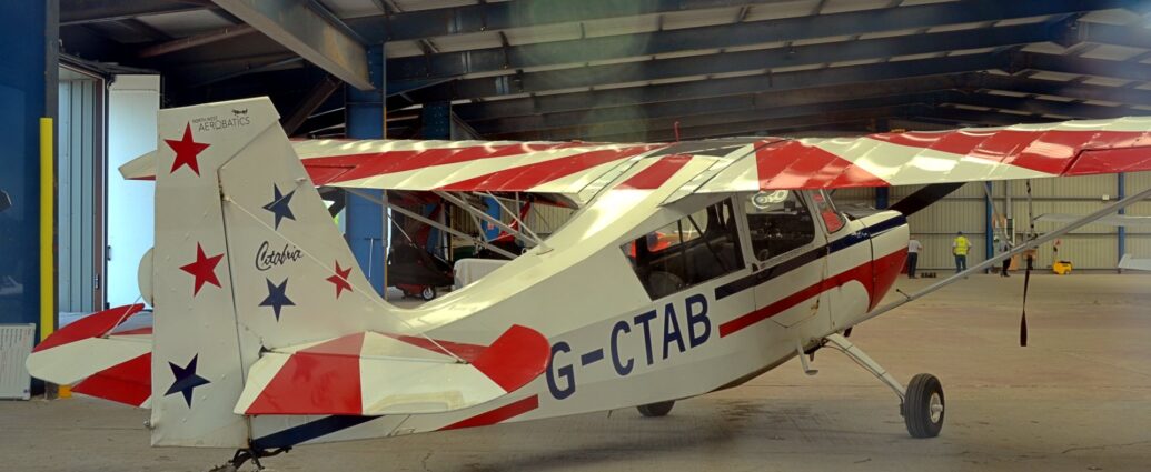 Rear-side view of a high-winged, single-engined light aircraft parked in a vaulted-roofed metal hangar. The plane is largely white, with red and blue stripes running along the body, overlaid with the black registration "G-CTAB" on the side of the rear fuselage. A red and white sun-burst pattern covers the top of both the wings and the horizontal stabiliser. The tail is white, with 3 blue and 3 red 5-pointed stars forming a rough triangle, the word "Citabria" in an ornate script in the middle, and the small text "North West Aerobatics right at the top. Grey brushed concrete floor fills the foreground and a lot of the background, with blue internal beams supporting metal walls and ceiling. A couple of people, one wearing a yellow hi-viz vest, are facing the far wall at the end. Blooms of green and bloom light are visible in the middle and edges of the frame, caused by shooting through slightly murky glass.