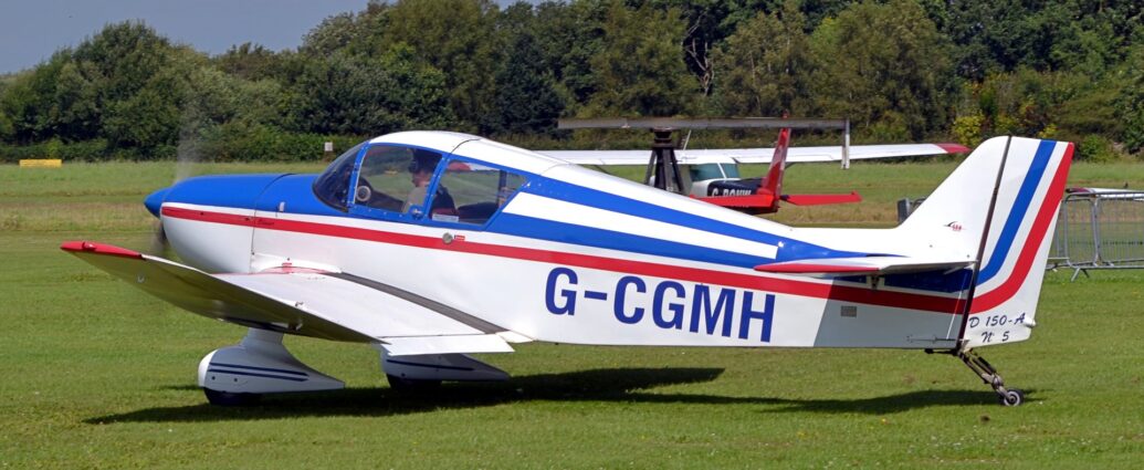 Side view of a white, single-engined light aircraft taxiing from right to left on a grass airfield. There is a blue and red stripe running along the body, with the blue registration "G-CGMH" on the lower rear fuselage. The outer panels on the wings are tilted slightly upwards, with the tips painted red. There are white spats covering the fixed main wheels under the wings, with a pair of thin blue stripes running along the sides. Green grass fills most of the frame, with a high-winged, single engined light aircraft parked just behind this one. Trees stretch across the frame in the distance, under grey-blue skies.