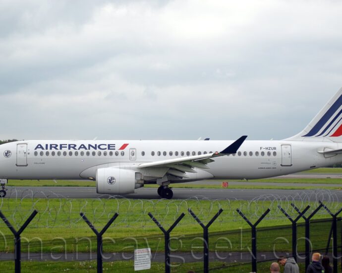 Side view of a twin engined jet airliner taxiing from left to right along a black taxiway. The plane is largely white, with dark blue "Air France" titles just ahead of a short, diagonal red band on the upper forward fuselage. The black registration "F-HZUR" is on the upper rear fuselage, with the last 2 letters repeated on the front of the open nose-wheel doors. There is additional black text "Airbus A220-300" on the lower rear fuselage. The white tail has a series of diagonal blue bands, thicker by the leading edge and getting thinner as they move rearwards, with a thicker red stripe at the lower rear, as well as a small circle of gold stars of the top of the tails. There is the blue outline of a white circle, containing a stylised image of a winged blue sea-horse, in the middle of the white engine pods, repeated on the side of the forward fuselage, in between the cockpit windows and the forward door. The up-turned wing-tips are entirely dark blue. A black mesh fence topped with razor-wire stretches across the bottom of the frame, with a small group of people on this side of the fence. Grass leads up to the taxiway the plane is on, with more grass and taxiways in the background, with the rear fuselage of a white, red, and black airliner on a runway facing out of the right of the frame in the distance. Rows of trees beyond that meet the cloudy grey sky.
