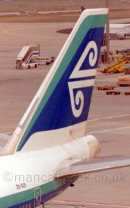 Vertically-oriented close-up of the rear fuselage and tail of a very large jet airliner. The plane is largely white, with a thick green and blue stripe running along the body, and the black registration "ZK-SUI" on the side of the upper rear fuselage. A green stripe fills the front and top of the white tail, with a large blue blob in the middle containing stylised white wind and waves. Grey concrete apron fills the background, with scattered airport furniture, things like yellow baggage carts or luggage loaders, or white mobile airstairs and vans, mostly on the right of the frame. A white, twin engined jet airliner with a dark blue tail is taxiing out of frame in the top right corner. Grass fills the top of the frame.