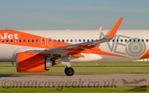 Close-up of the wing and centre fuselage of a twin engined jet airliner parked on a grey concrete runway facing to the left. The plane is mostly white, with a wide, diagonal orange band running from the top of the fuselage ahead of the wing, to the belly just aft of the wing, getting narrower as it moves rearwards.# The white word "Jet" can be seen at the top left of the stripe. Another wide, diagonal orange stripe starts on the lower rear fuselage reaching towards the tail. Above and between the two orange stripes is the very large, grey word "Neo", with the last letter resembling the front of a jet engine, showing off the fan blades. The engines, mounted under and slightly forward of the wing, are the same bright orange, as are the flap track covers under the rear of the wing, and the large, up-turned wingtips. In the foreground, lush green grass lines both sides of the runway, as well as a taxiway visible under the body. Above the plane, blue sky with hints of grey cloud fill the rest of the frame.