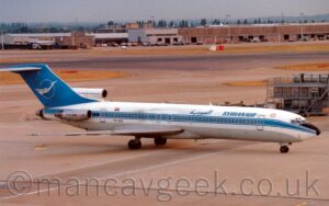 Side view of a three engined jet airliner with the engines mounted on the sides and top of the rear fuselage, taxiing from left to right along a grey concrete taxiway. The plane is largely white, with a blue stripe running along the body, covering the passenger cabin windows, with thin blue pinstripes above and below, a grey belly, and blue "SyrianAir" titles on the upper forward fuselage, in English at the front, and in an Arabic script just forward of the wings. The black registration "YK-AGA" is on the side of the lower rear fuselage, with a small, red, white, and black Syrian flag above the windows just above it. The bare metal engine pods mounted on the sides of the rear fuselage have black text "Boeing 727" on the sides. The centre engine pod, in front of and at the base of the tail, is white, with some blue text in an Arabic script. The tail is blue, with the thin outline of a white circle, containing a stylised image of a white flying bird. Grey concrete apron fills the foreground, with a grey metal storage rack for luggage containers in the background on the right of the frame, and more taxiways and a runway, separated by yellow grass, filling the rest. Grey and brown cargo sheds stretch across the frame in the distance, with trees beyond, under grey sky.