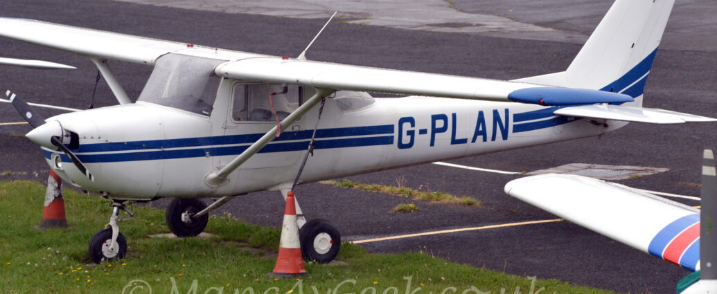 Slightly elevated side view of a high-winged, single-engined light aircraft parked facing to the left on small concrete pads surrounded by grass. The plane is largely white, with a pair of blue stripes running along the body, overlaid by the blue registration "G-PLAN" on the sides of the rear fuselage. A pair of orange and white traffic cones sit on the grass, one on either side, attached by long cables to the top of the white support struts that run from the bottom of the fuselage to the wings. Green grass fills the foreground, with the nose of another light aircraft peeking into frame in the bottom right corner. Black asphalt apron fills the background.
