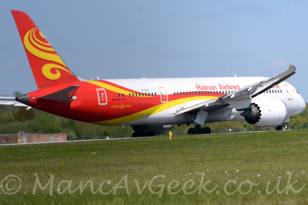 Side view of a twin engined jet airliner moving from left to righ along a runway. The plane is mostly white, with a red and yellow stripe running along the body, spilling over the front of the wings, and running up and down to fill the rear fuselage, with a second yel;low stripe on the upper rear. The tail is red, with yellow swirls in the middle. There are red "Hainan Airlines" titles on the upper forward fuselage, just aft of the forward mid-cabin door, with the same titles in a Chinese script further forward, partially obscured by the wing. There is black text "Boeing 787-8" under the rear-most cabin windows, above larger, yellow "DreamLiner" text. The black registration "B-2731" si on the upper rear fuselage, with the number "2731" repeated on the sides of the open nose-wheel doors. White engine pods, with scalloped rear edges, are suspended under the wings. Green grass fills the foreground, with trees in the background, those on the left blurred by the jellyfied hot air coming from the rear of the engines. Blue sky fills the rest of the frame.