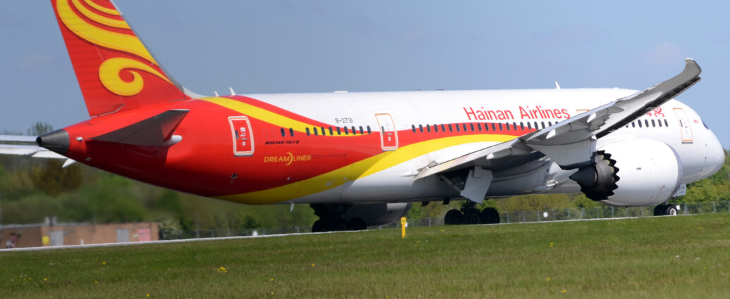 Side view of a twin engined jet airliner moving from left to righ along a runway. The plane is mostly white, with a red and yellow stripe running along the body, spilling over the front of the wings, and running up and down to fill the rear fuselage, with a second yel;low stripe on the upper rear. The tail is red, with yellow swirls in the middle. There are red "Hainan Airlines" titles on the upper forward fuselage, just aft of the forward mid-cabin door, with the same titles in a Chinese script further forward, partially obscured by the wing. There is black text "Boeing 787-8" under the rear-most cabin windows, above larger, yellow "DreamLiner" text. The black registration "B-2731" si on the upper rear fuselage, with the number "2731" repeated on the sides of the open nose-wheel doors. White engine pods, with scalloped rear edges, are suspended under the wings. Green grass fills the foreground, with trees in the background, those on the left blurred by the jellyfied hot air coming from the rear of the engines. Blue sky fills the rest of the frame.