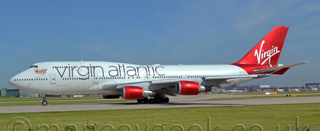 Side view of a very large, 4 engined jet airliner lining up on a grey runway as it prepares for take-off. The plane is largely white, with large, thin, purple "Virgin Atlantic" titles on the upper forward fuselage, mostly between the windows of the upper and lower decks. There is an image of a flying woman, wearing a red leotard, and wearing a UK flag as a cape, under the cockpit windows at the front of the upper deck. The partial registration "-VAST" can be seen on the lower rear fuselage, the last two letters "ST" also appearing on the open nose-wheel doors. The tail and rear fuselage are a shiny, scarlet redd, with seemingly hand-drawn, white "Virgin" titles across the middle, and the small white letters "ST" right at the top. The engine pods are a similar shiny scarlet, as are the up-turned wing-tips. Green grass fills the foreground, with a couple of bushes in the bottom left corner, and black metal mesh fence supported by black metal poles runs across the bottom of the frame. More grass is visible in the background, under the body of this plane, with a terminal building on the right, with several planes parked against it. Pale blue sky with wisps of white cloud fill the top of the frame.