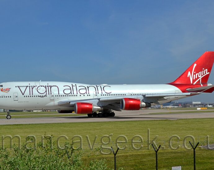 Side view of a very large, 4 engined jet airliner lining up on a grey runway as it prepares for take-off. The plane is largely white, with large, thin, purple "Virgin Atlantic" titles on the upper forward fuselage, mostly between the windows of the upper and lower decks. There is an image of a flying woman, wearing a red leotard, and wearing a UK flag as a cape, under the cockpit windows at the front of the upper deck. The partial registration "-VAST" can be seen on the lower rear fuselage, the last two letters "ST" also appearing on the open nose-wheel doors. The tail and rear fuselage are a shiny, scarlet redd, with seemingly hand-drawn, white "Virgin" titles across the middle, and the small white letters "ST" right at the top. The engine pods are a similar shiny scarlet, as are the up-turned wing-tips. Green grass fills the foreground, with a couple of bushes in the bottom left corner, and black metal mesh fence supported by black metal poles runs across the bottom of the frame. More grass is visible in the background, under the body of this plane, with a terminal building on the right, with several planes parked against it. Pale blue sky with wisps of white cloud fill the top of the frame.