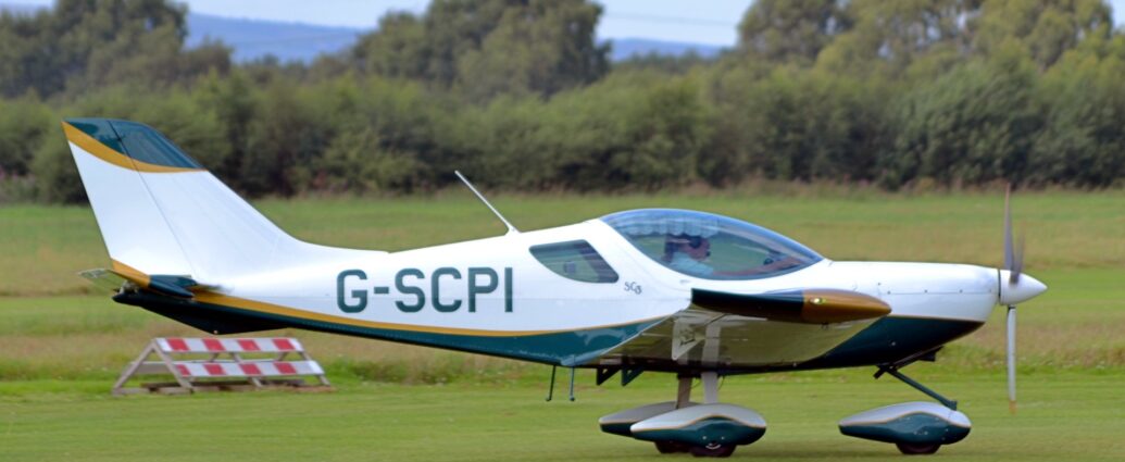 Side view of a white, single engined light aircraft taxiing from left to right on a grass airfield. The plane is white on top, with a green belly, with a gold stripe separating the 2 colours. The same colour scheme is repeated on the spats covering the wheels on the fixed undercarriage. The ends of the wings are gold at the front and green ant the back. Green grass fills most of the frame, leading up to green grass in the distance, under grey skies.