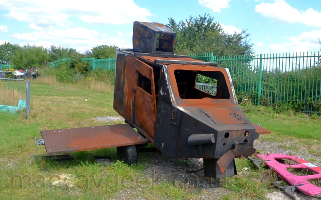 A rather weather-beaten and rusty lump of metal that has been left on grass, ostensibly to rot. Vaguely resembling a light aircraft designed by someone who had only heard a description of an aeroplane, there is a place in the nose for an piston engine, with a cockpit just after that, and what appears to be a single vestigial jet engine on the top rear of the fuselage. The whole thing is covered in rust except where it has been burned, mostly around the nose and the wings. It is surrounded by grass, with a red plastic barrier lying on the ground beside it, and a green metal fence off to one side of the background, with bushes beyond that. Blue sky with fluffs of white cloud fill the rest of the frame.