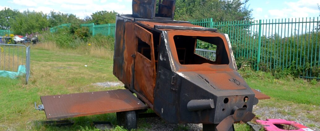 A rather weather-beaten and rusty lump of metal that has been left on grass, ostensibly to rot. Vaguely resembling a light aircraft designed by someone who had only heard a description of an aeroplane, there is a place in the nose for an piston engine, with a cockpit just after that, and what appears to be a single vestigial jet engine on the top rear of the fuselage. The whole thing is covered in rust except where it has been burned, mostly around the nose and the wings. It is surrounded by grass, with a red plastic barrier lying on the ground beside it, and a green metal fence off to one side of the background, with bushes beyond that. Blue sky with fluffs of white cloud fill the rest of the frame.