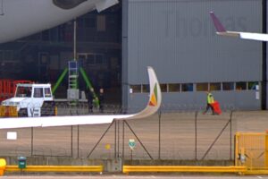 Close-up of the up-turned, curved right wingtip of a jet airliner parked out of frame on the left, facing away from the camera, with the wing stretching into frame from the left. The wing is almost entirely white, with green, yellow, and red wedges on the vertical face of the wingtip. The purple, up-turned wingtip of another plane is visible towards the top of the frame on the right, in front of the large open door of a grey maintenance hangar, with part of the rear fuselage of an even larger plane visible inside the hangar. A man wearing a yellow hi-viz vest can be seen dragging a red rubbish bin in front of the hangar.